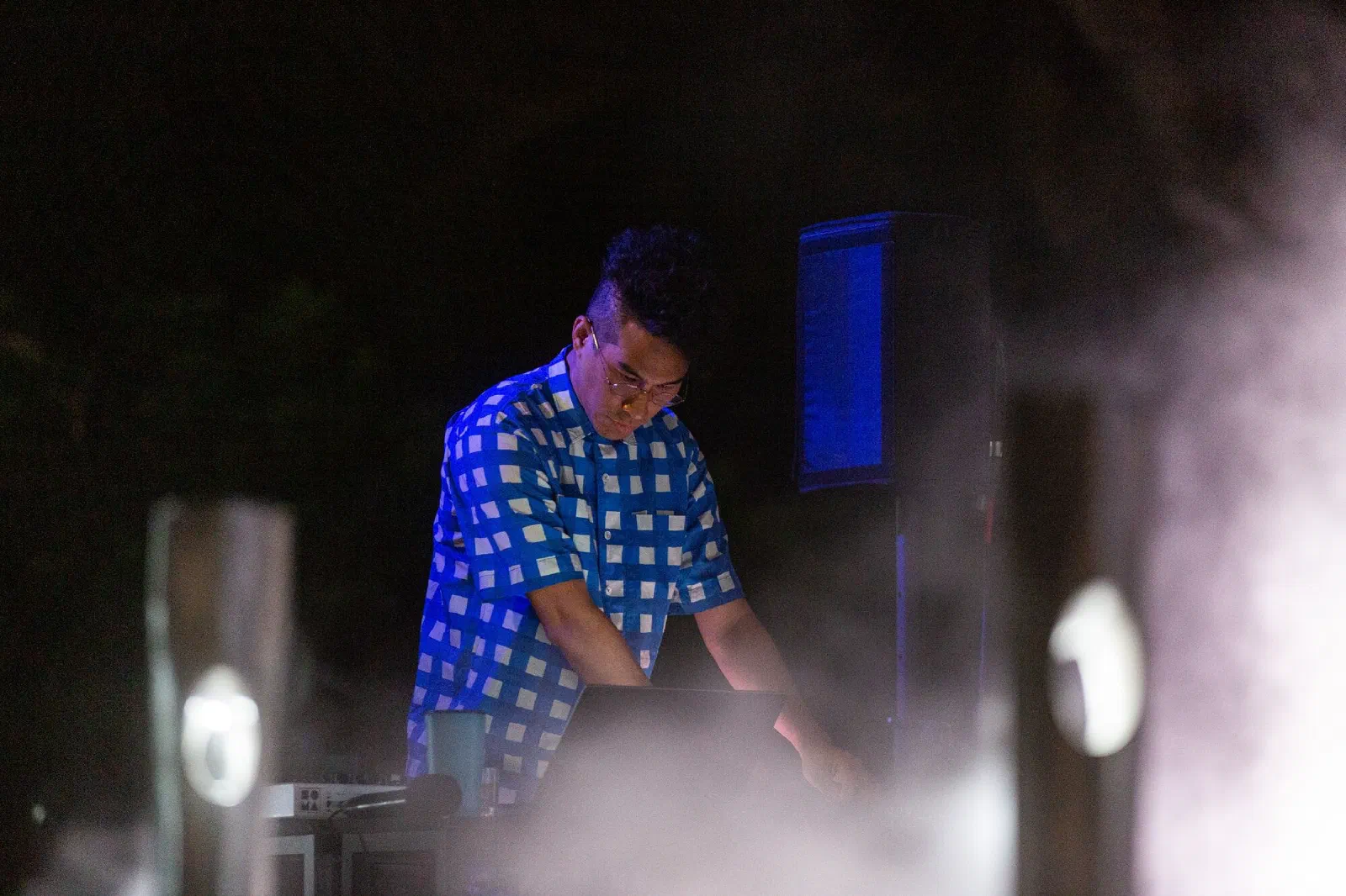 A person in a blue checked shirt works at a laptop and equipment table in blue light, with a speaker behind and mist in the foreground.