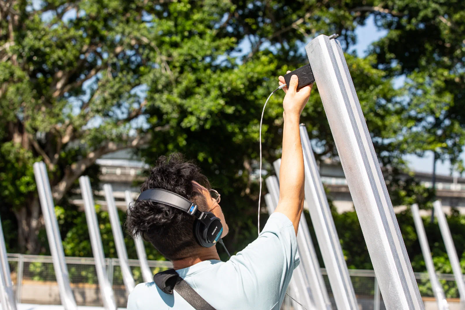 A person in headphones holds a small microphone against a tall aluminium pole, with trees and other poles behind.