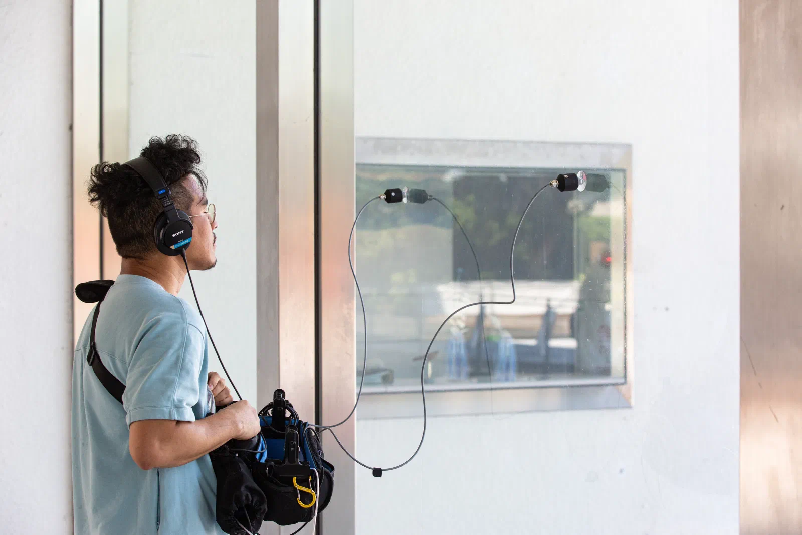 A person in headphones stands beside a window, with two contact microphones on suction cups attached to the glass and connected to a recorder bag.