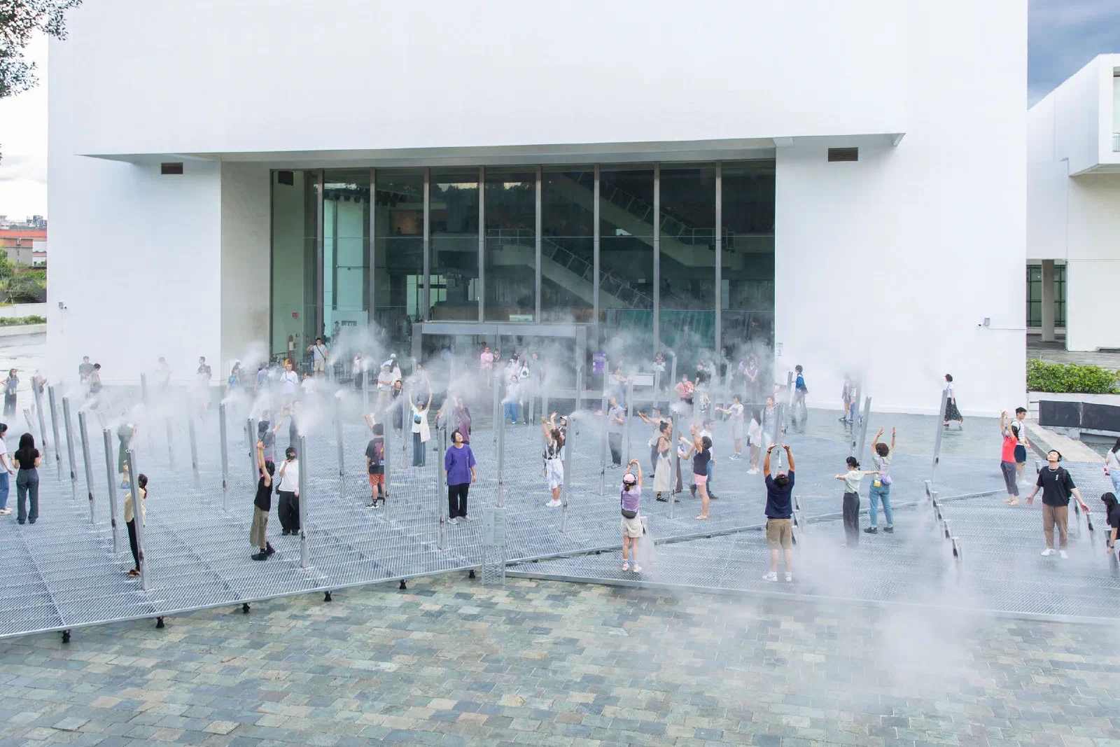 Participants stand among tall poles releasing mist on a grated platform outside a white museum building, many with arms raised toward the mist.