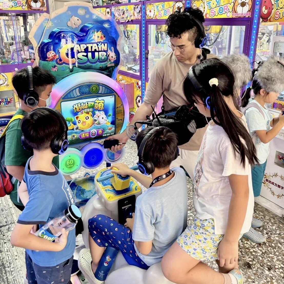 In an arcade, several children in headphones gather around a submarine arcade machine while a facilitator holds a shotgun microphone toward it.