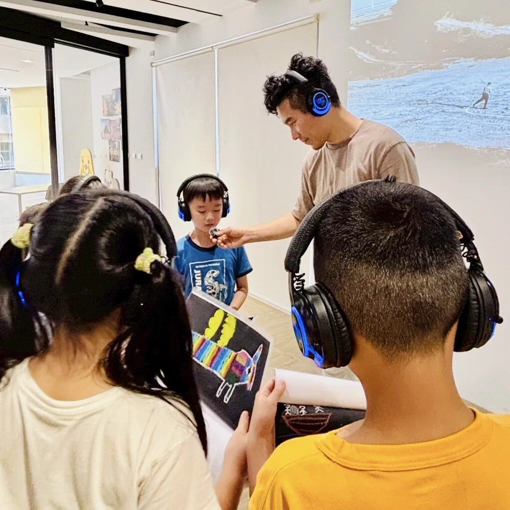 A facilitator holds a small microphone toward a boy in headphones, while two children in the foreground hold up their drawings of colourful creatures.