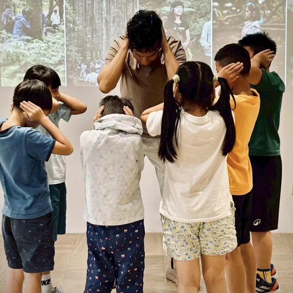 A facilitator and six children stand in a semicircle facing a wall, each with both hands cupped over their ears, in front of projected forest photographs.