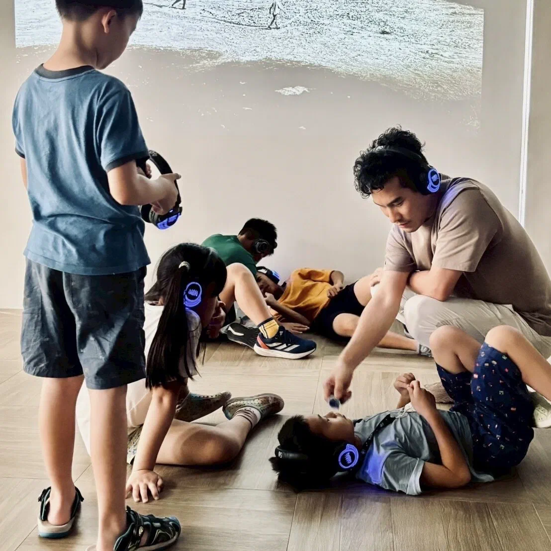 Children in blue-lit headphones lie and crouch on a wooden floor while a facilitator kneels beside them, with a projected shoreline image on the wall behind.