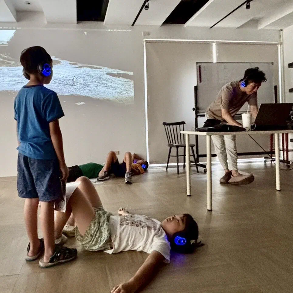 A wide view of a bright room where children in headphones lie on the floor and one boy stands, a projected shoreline on the wall and a facilitator at a laptop on a table.