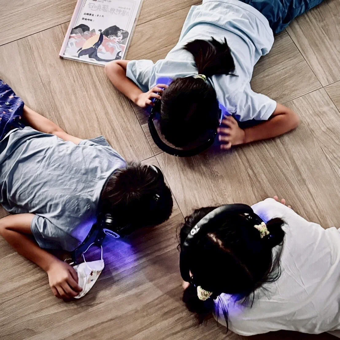 Seen from above, three children lie on their fronts on a wooden floor wearing headphones that glow purple, with a printed programme booklet beside them.