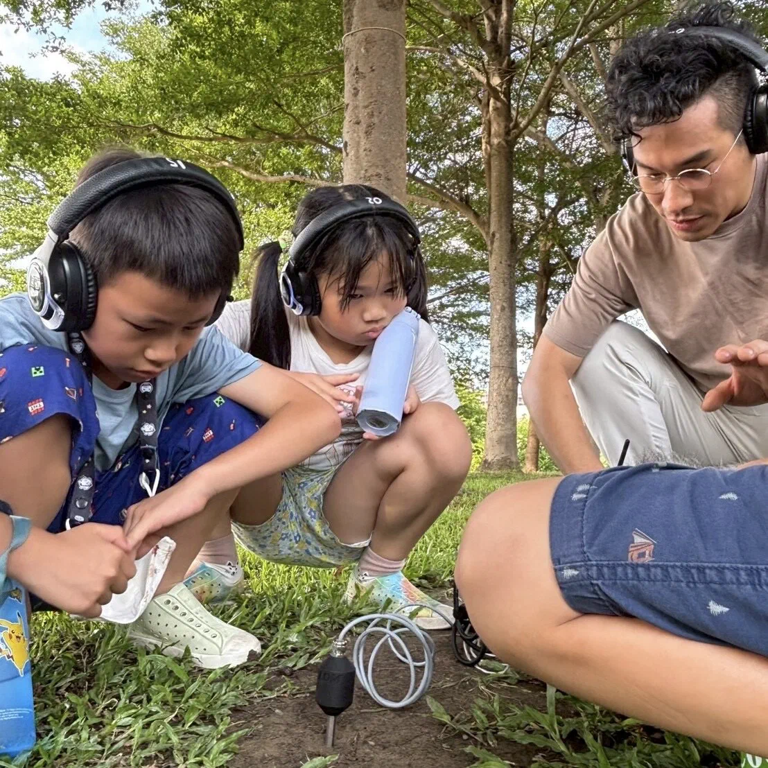 Two children in headphones crouch on grass beside a small microphone pushed into the soil, one holding a blue tube to her ear, while the facilitator watches.