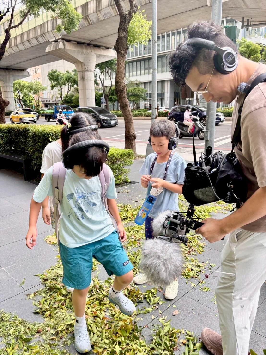 On a city pavement covered in cut leaves, a child steps on the leaves while another child and a facilitator with a furry windshielded microphone listen, with a raised road behind.