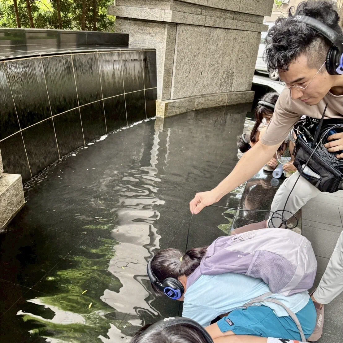 Beside a black stone water feature, a facilitator in headphones holds a cable out over the water while a child in headphones leans in to listen, with a recorder bag at his side.