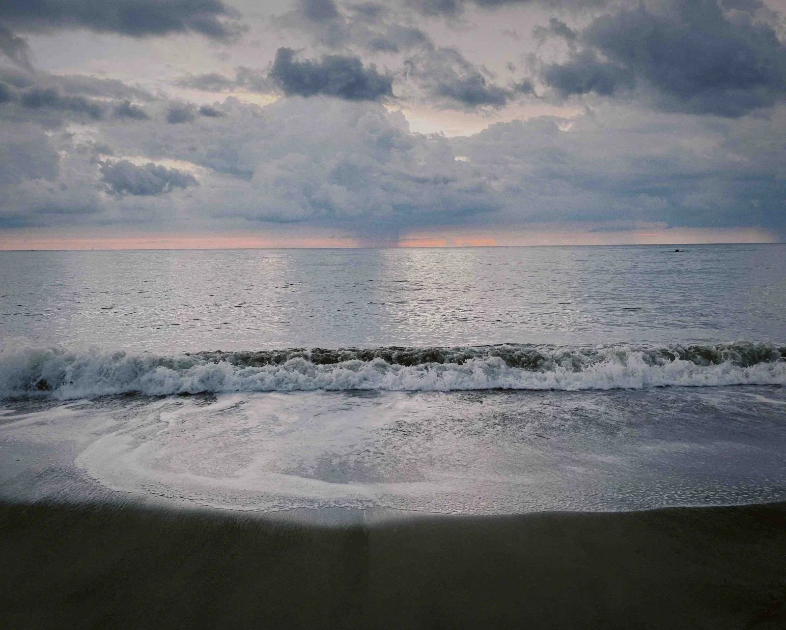 A wave breaks and washes up a dark sand beach under heavy clouds, with a band of orange light on the horizon.