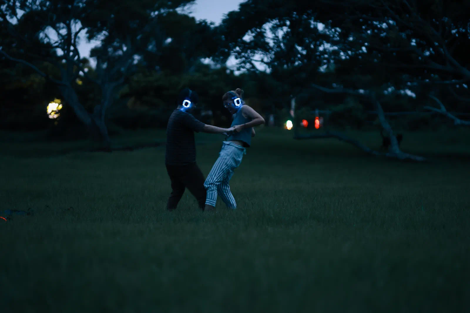 Two participants in glowing headphones hold hands and dance on dark grass at dusk.