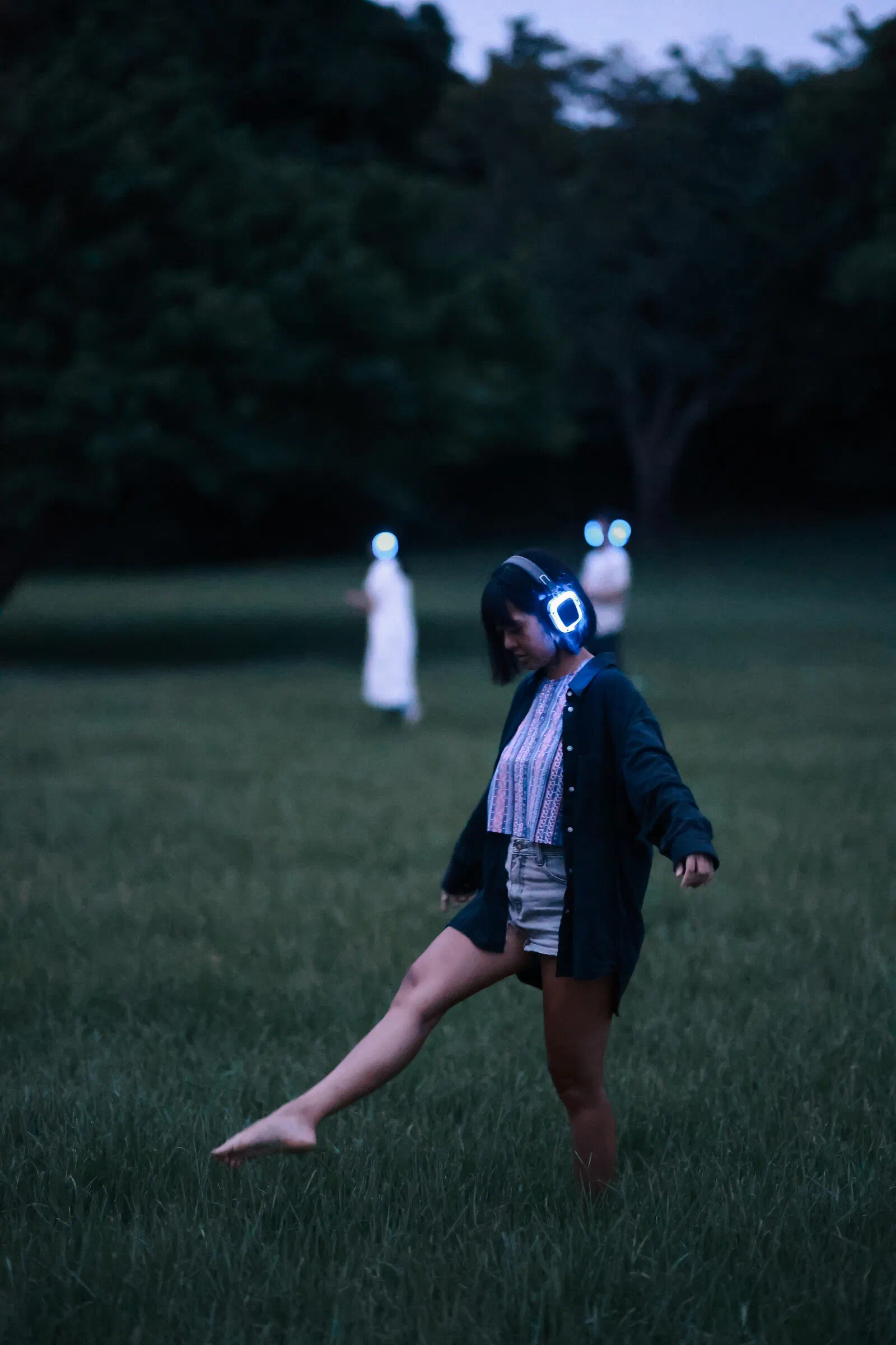 A barefoot participant in glowing headphones lifts one leg mid-step on the grass at dusk, with two blurred figures behind.