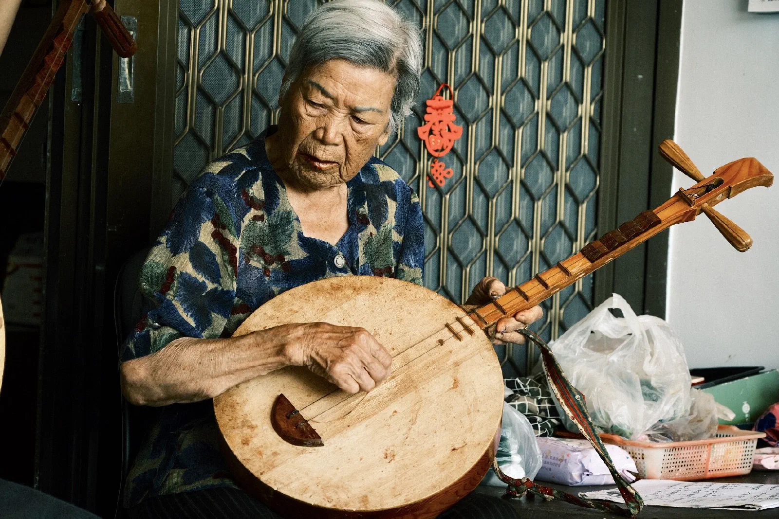 An elderly woman plays a wooden moon lute, looking down at the strings, in a doorway with a patterned metal screen door behind her.