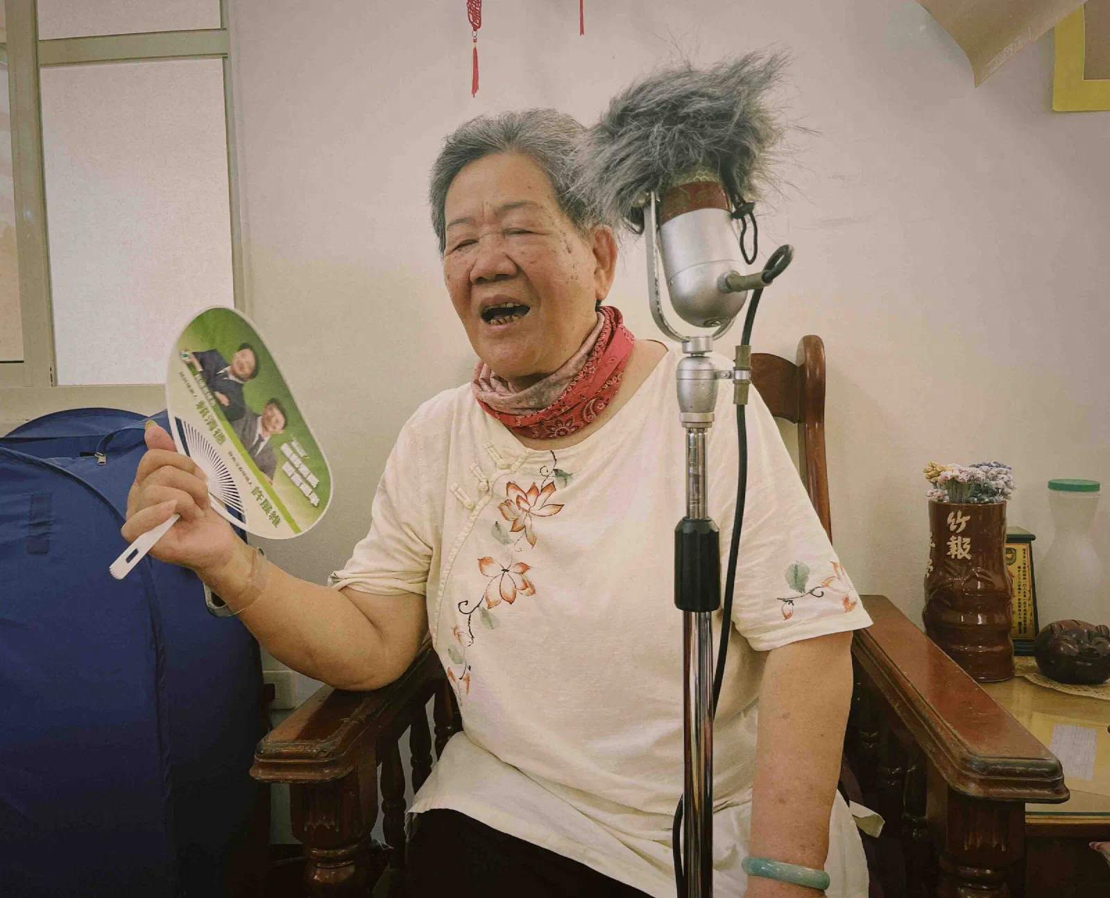An elderly woman sings with her mouth open, holding a paper fan, beside a vintage microphone in a fur windshield.