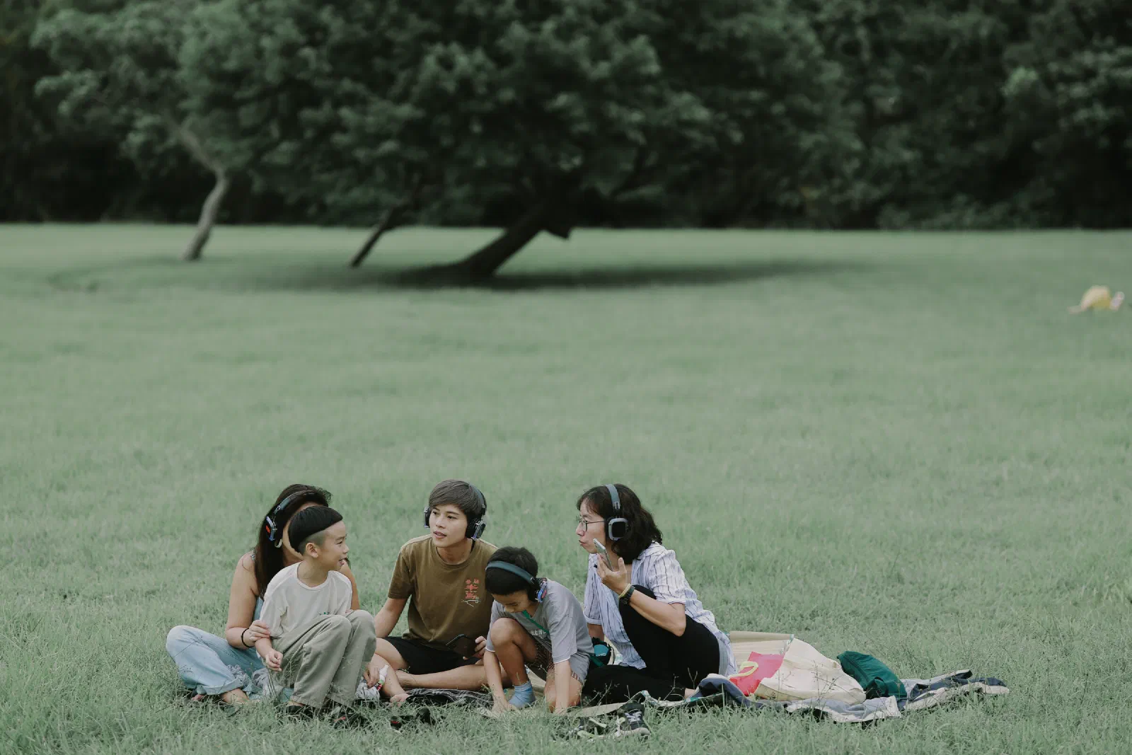 A family group sits together on a blanket on a wide lawn, wearing headphones and talking, with a leaning tree behind.