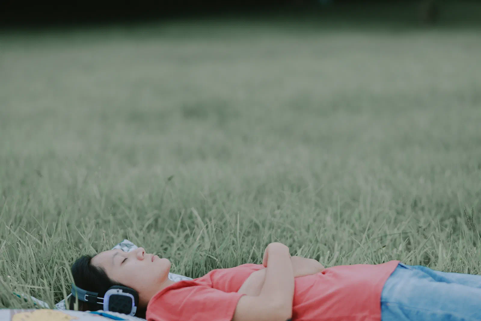 A person lies on their back in tall grass with arms crossed over their chest, eyes closed, wearing headphones.