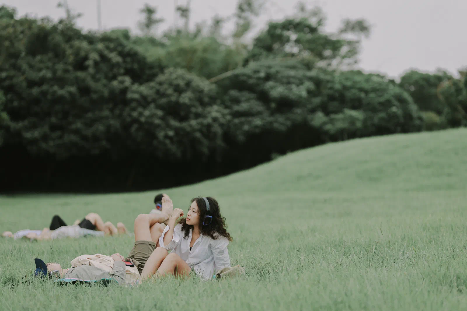 A woman sits in a meadow wearing headphones and looking to the side, while other participants lie in the grass behind her.