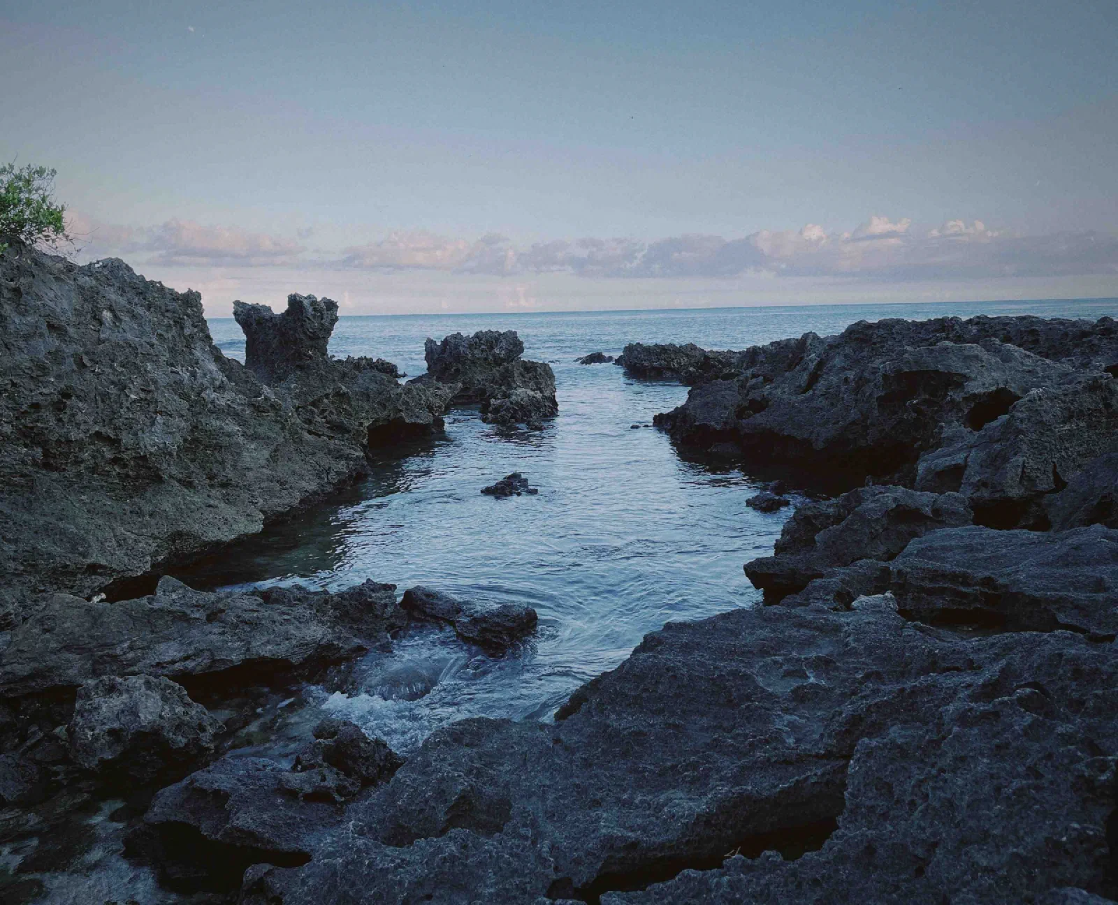 Dark, weathered coastal rocks frame a calm tidal pool that opens to the sea under a pale evening sky.
