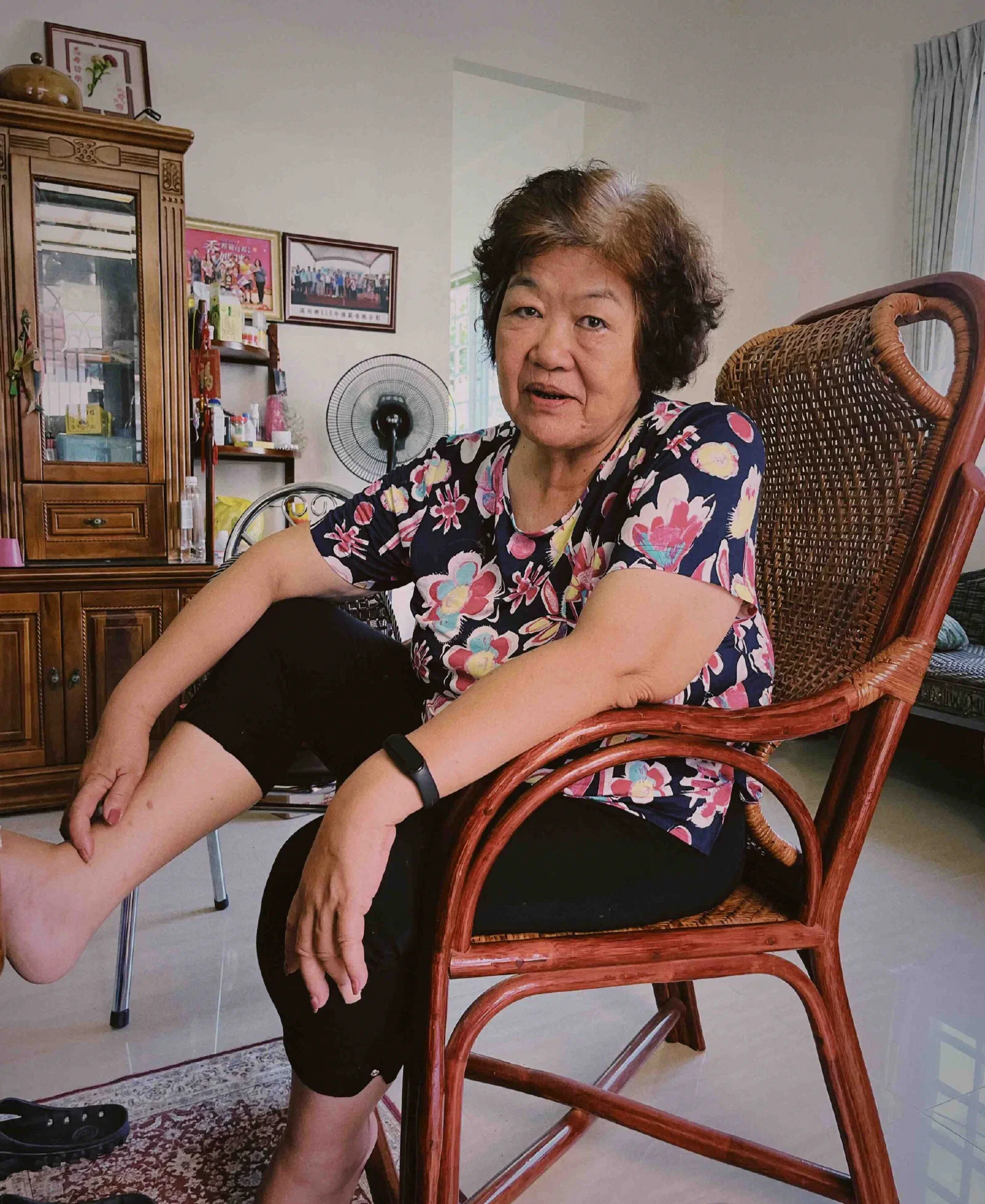 A woman sits sideways in a rattan chair in a living room, resting one arm over the back and speaking toward the camera.