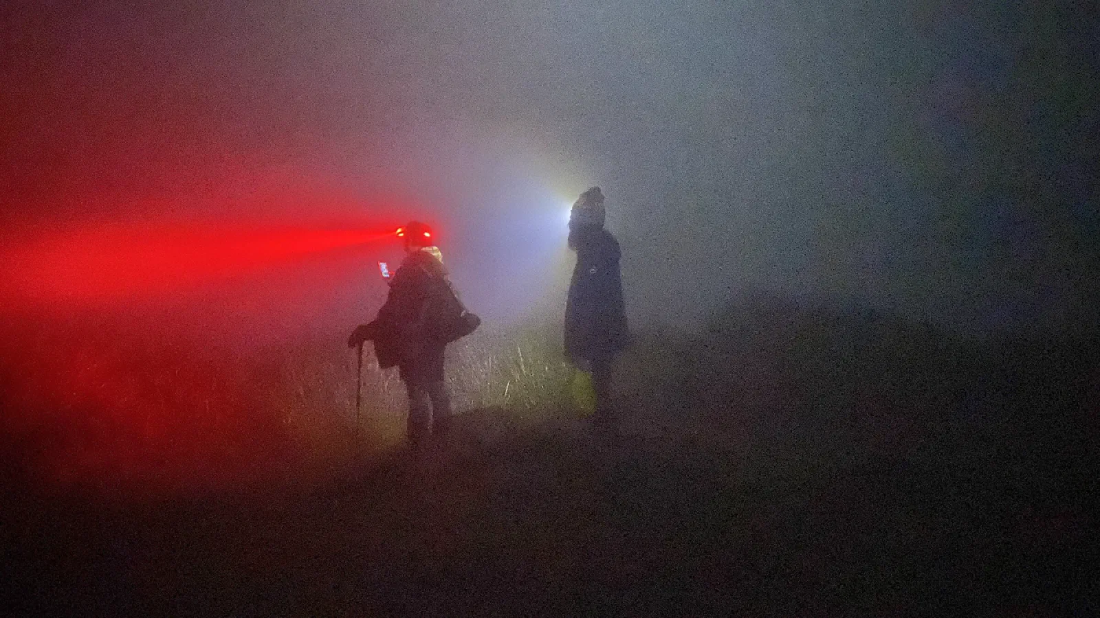 Two people with headlamps, one shining red and one white, stand in thick night fog on a grassy ridge, seen only as silhouettes.