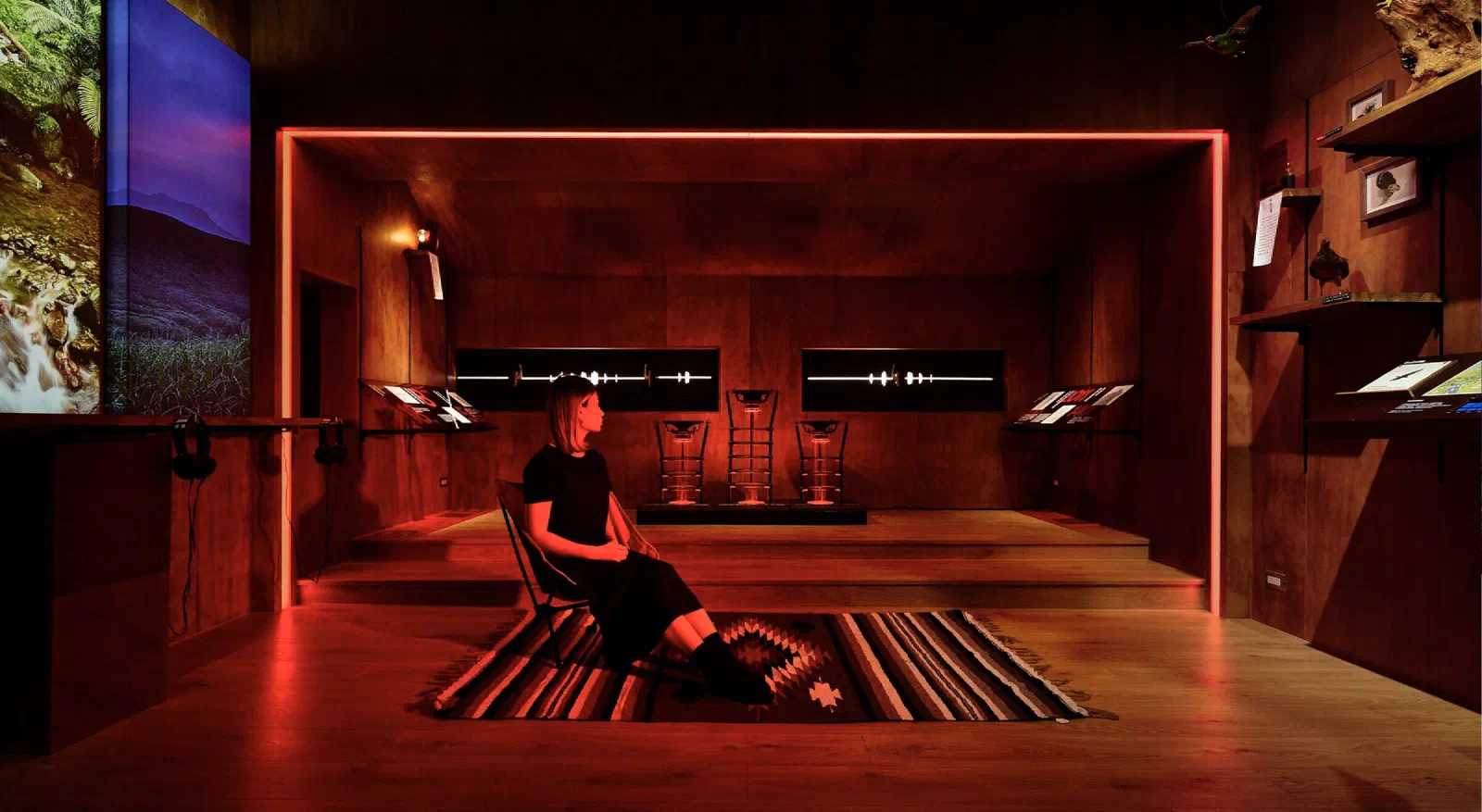 A woman sits on a folding chair on a woven rug in a dark, red-lit wooden room, facing three speakers on a low stage and two glowing waveform displays, with mountain landscape panels at the left and framed specimens on shelves at the right.