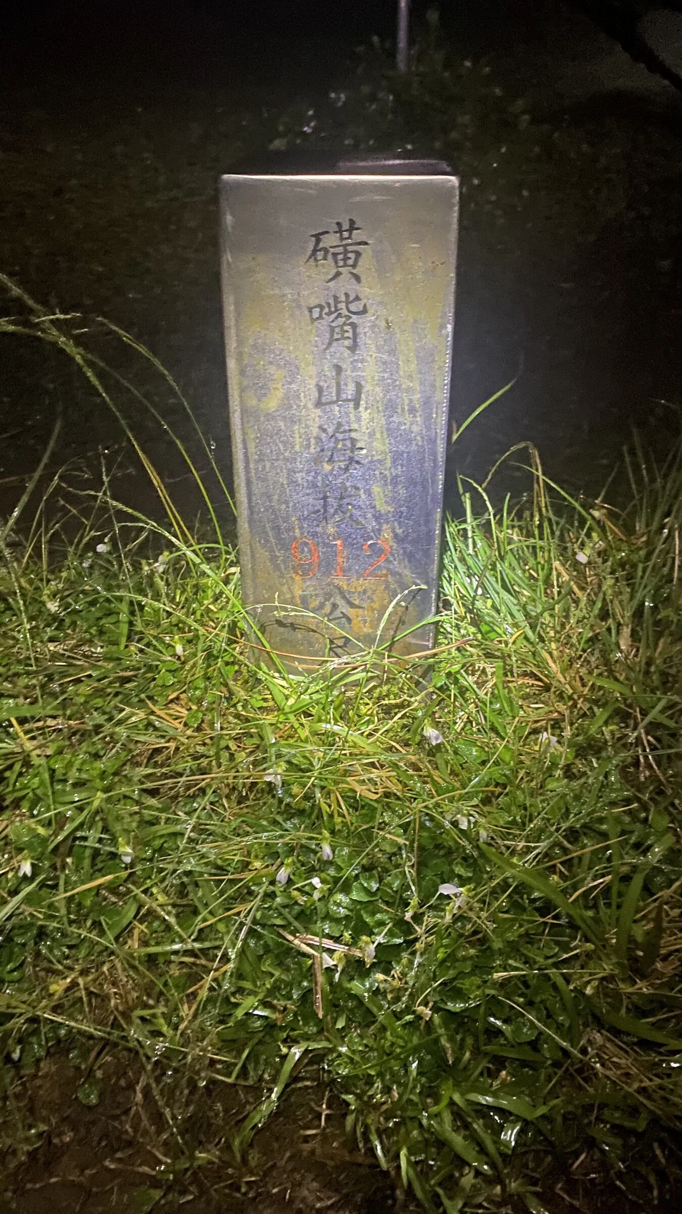 At night, a weathered stone marker inscribed with a mountain name and an altitude of 912 metres stands in wet grass, lit by a torch.